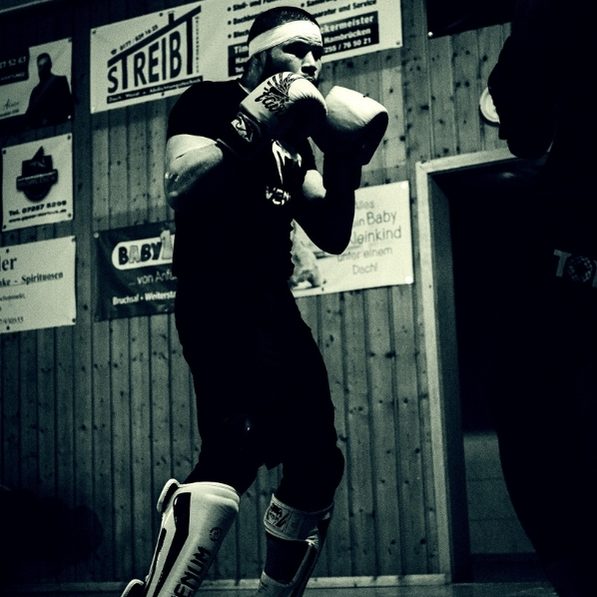 Ein Wettkämpfer trainiert mit Boxhandschuhen und Schienbeinschoner in einem schlichten Trainingsraum.